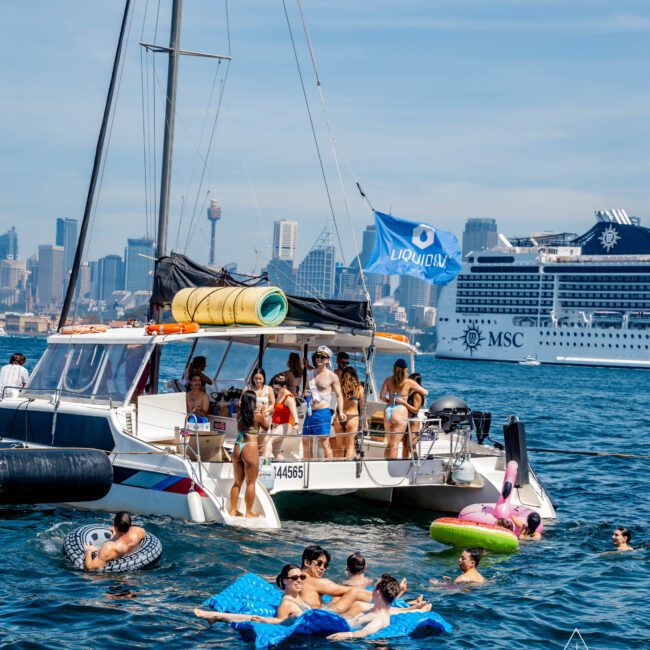 A group of people enjoying a sunny day on a yacht and in the water. Some are lounging on inflatable floats, while others stand on the boat. The city skyline and a large cruise ship are visible in the background.