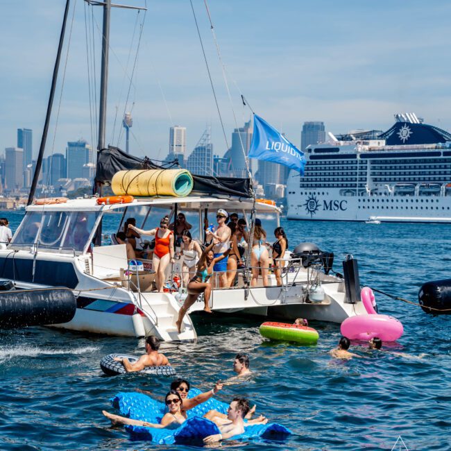 A group of people are enjoying a sunny day on a boat and in the water. Some are on inflatable floats. In the background, a large cruise ship is visible. The skyline of a city can be seen in the distance.