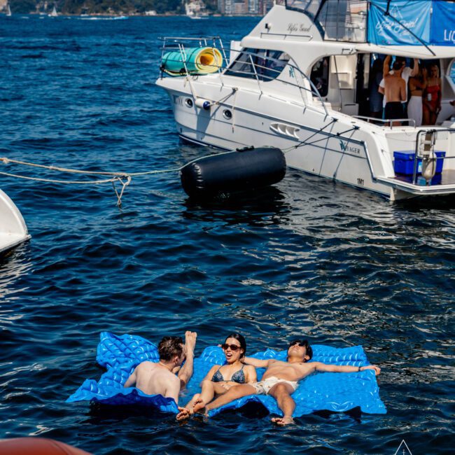 Three people relax on blue floating mats in the water near a yacht. The yacht has people on board, with city buildings visible in the background across the water. The scene is bright and sunny.