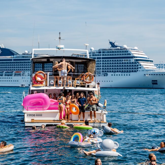 A crowded boat with people in swimsuits, some sitting, others diving. Bright inflatables, including a pink flamingo and a rainbow, float nearby. A large cruise ship is visible in the background against a clear blue sky.