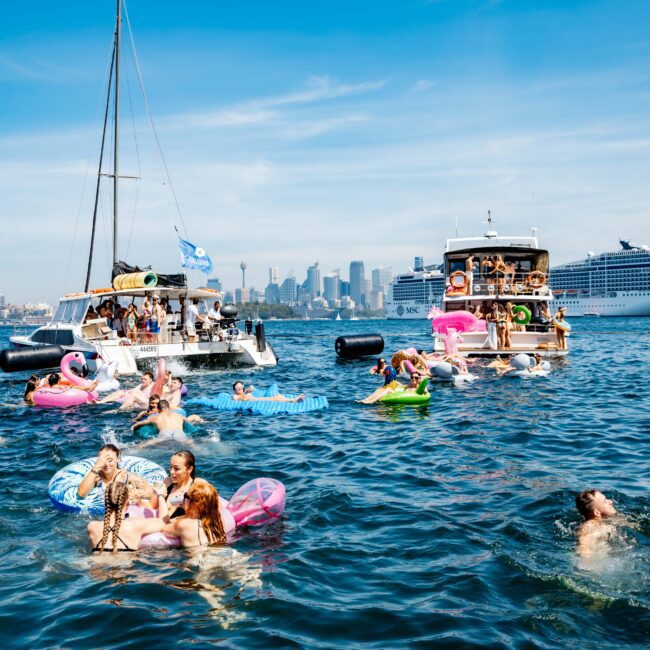 People swimming and relaxing on inflatable rings in a bustling harbor, surrounded by boats. A cruise ship and city skyline are visible in the background under a clear blue sky.