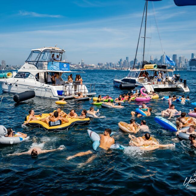 A large group of people enjoying a sunny day on the water, floating on colorful inflatable rafts near several boats. The city skyline is visible in the background under a clear blue sky.