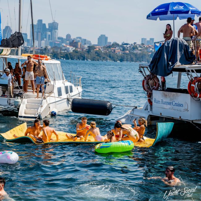People are enjoying a sunny day on a boat party. They are relaxing on floating mats and tubes in the water between a catamaran and a motorboat named "Rum Runner Cruises." The city skyline is visible in the background.