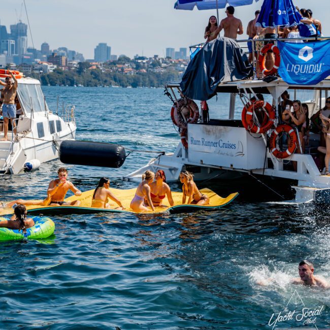People enjoy a sunny day on the water, with a group on a yellow inflatable connected to a boat labeled "Rum Runner Cruises." Others swim nearby, with city buildings visible in the background. The scene is lively with a clear blue sky.