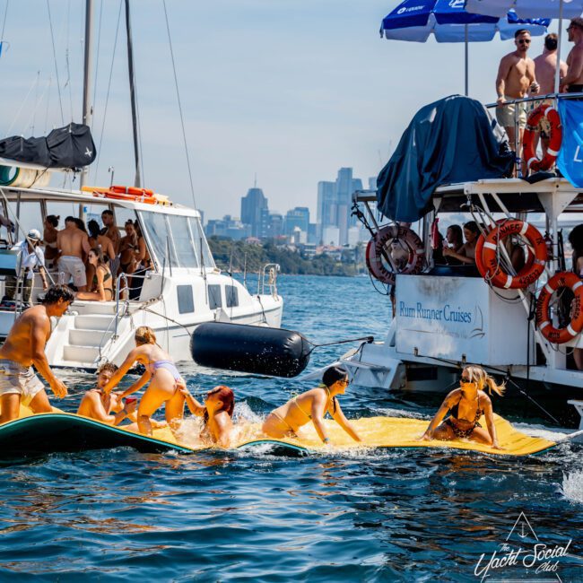 People enjoying a sunny day on the water, swimming and relaxing on boats. A group is on a large inflatable mat. One boat has "Rum Runner Cruises" on it. The city skyline is visible in the background.