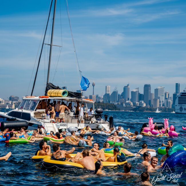 A lively group of people floats on colorful inflatables near anchored boats. The city skyline is visible in the background under a clear blue sky. The water is calm, and the atmosphere is festive.