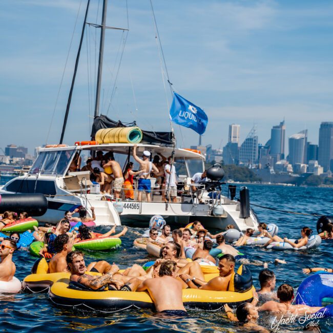 A large group of people on inflatable rafts and floats enjoy a sunny day on the water near a boat. A city skyline is visible in the background. The scene is lively, with people socializing and relaxing.