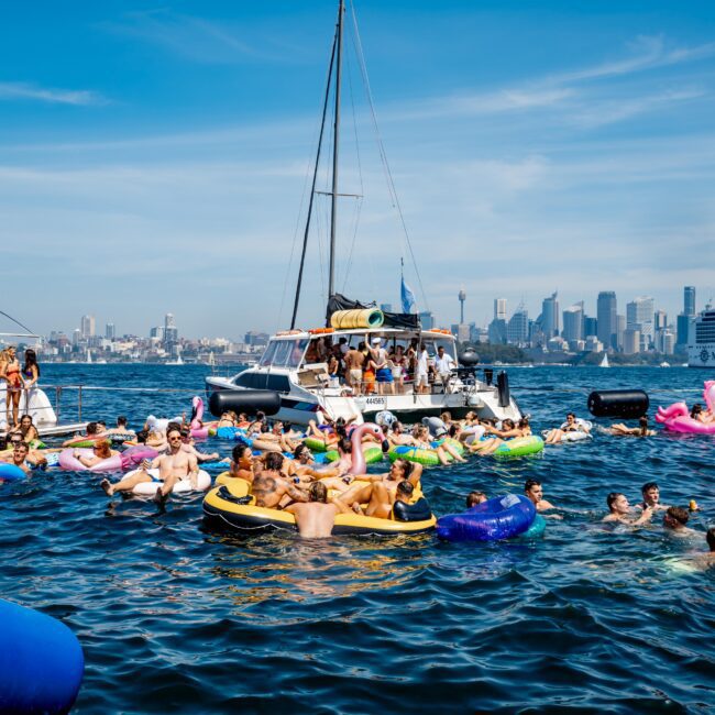 A vibrant scene on the water with several people on inflatable floats and boats, enjoying a sunny day. The city skyline, including the Sydney Tower, is visible in the background under a clear blue sky.