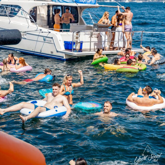 People enjoying a sunny day on the water, swimming and lounging on inflatable tubes near a yacht. Some are socializing on the boat's deck, and various inflatables surround the area. City skyline visible in the background.
