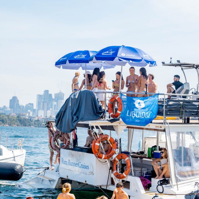 A group of people enjoying a sunny day on a boat, with several standing under umbrellas on the upper deck. Others are playing on a yellow floating mat next to the boat. The city skyline is visible in the background.