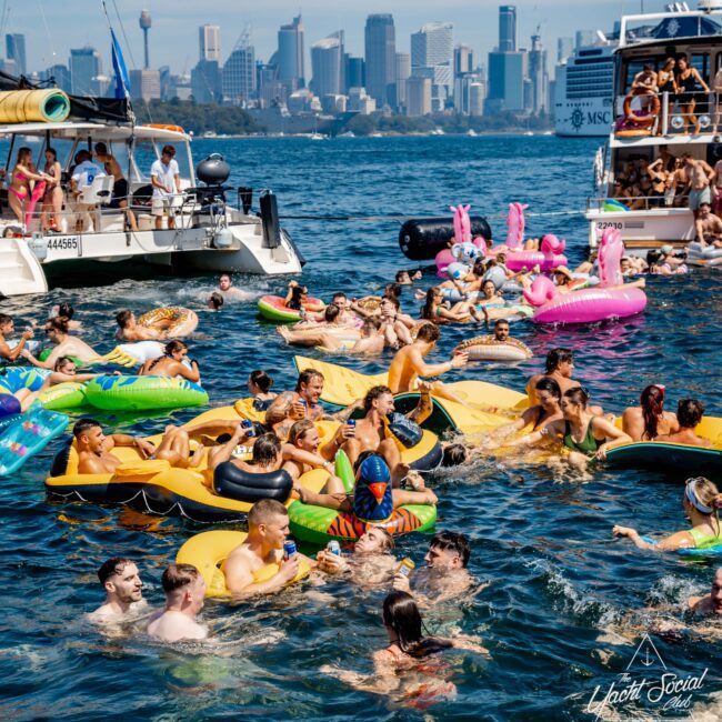 A lively group of people enjoying a party on inflatable floats in the water, surrounded by boats. The city skyline is visible in the background. Floats include colorful designs like pineapples and ducks, and people are socializing and swimming.