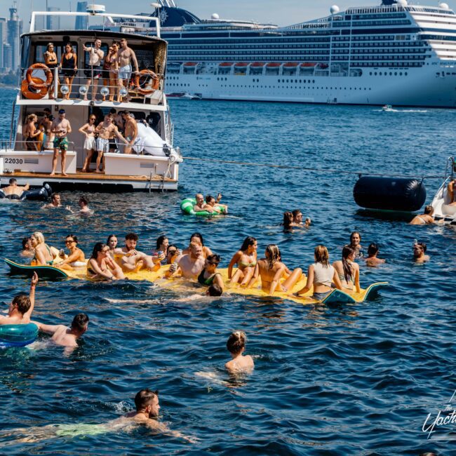 A lively scene of people enjoying a sunny day on the water. Many are swimming or lounging on colorful inflatable rafts. Boats and a large cruise ship are in the background. The atmosphere is festive and summery.