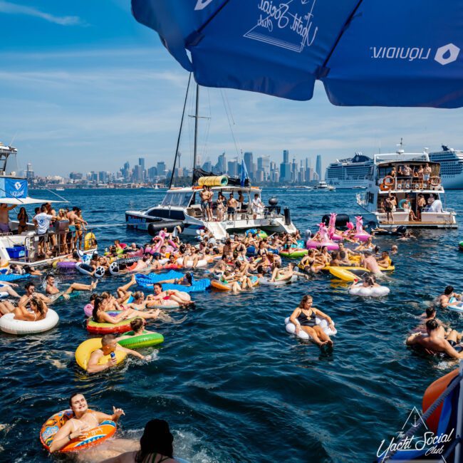 A large group of people enjoying a sunny day on the water, floating on various inflatable devices near boats. The city skyline is visible in the background. A blue umbrella and a "Yacht Social Club" logo are present.