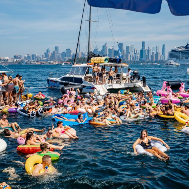 A lively scene of people enjoying a sunny day on water floats, with various colorful inflatables. Surrounding boats are filled with more people. A city skyline and cruise ship are visible in the background.