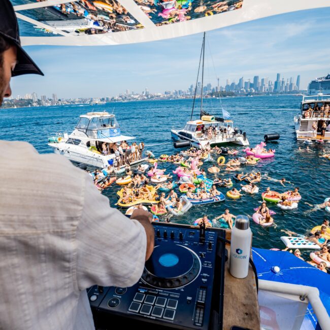 DJ playing on a yacht with a view of people on inflatables in water. The scene includes several boats and a city skyline in the background.