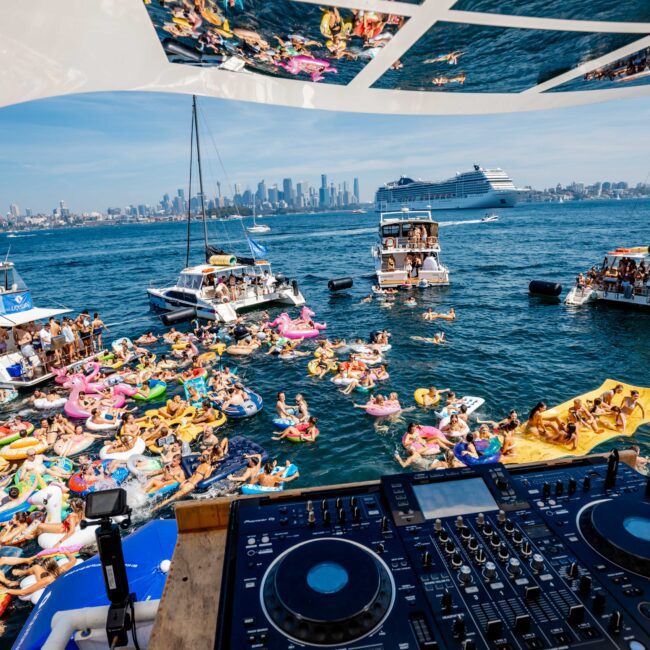 A vibrant boat party on the water with people on jet skis and colorful floaties. Boats are gathered together, and a DJ setup is in the foreground. The city skyline and a large cruise ship are visible in the background.