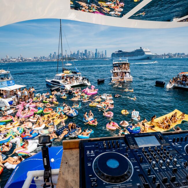 View from a DJ booth on a yacht party. People are floating on inflatable rafts in the water. The city skyline and a large cruise ship are in the background. Partygoers enjoy a sunny day on the water.