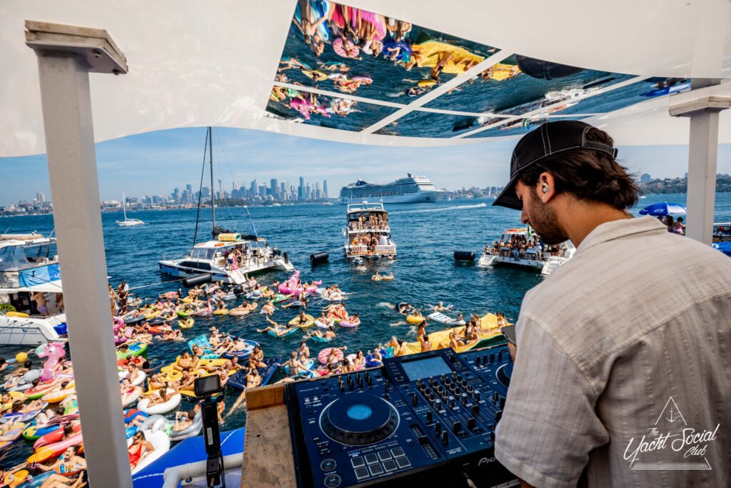 A DJ plays music on a boat surrounded by people on inflatables and boats in a vibrant sea party scene. The city skyline and a large cruise ship are visible in the background.