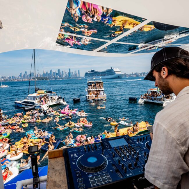 A DJ plays music on a boat surrounded by people on inflatables and boats in a vibrant sea party scene. The city skyline and a large cruise ship are visible in the background.