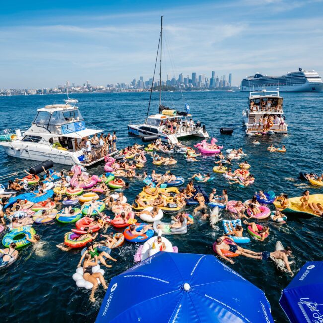 A lively scene of people on colorful inflatables in a harbor with several boats nearby. The city skyline is visible in the background, and there's a large cruise ship on the right. Two blue umbrellas are in the foreground.