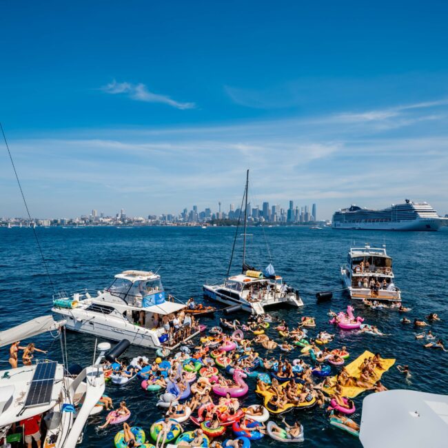 A large group of people enjoying a sunny day in a bay full of boats and colorful inflatables. The city skyline is visible in the background, and a cruise ship passes by on the right. The water is calm and the sky is clear.