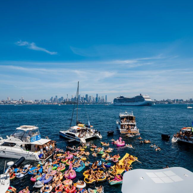 Numerous boats gather in a harbor with people lounging on colorful inflatables in the water. A large cruise ship is visible in the background, with a distant city skyline and clear blue sky overhead.