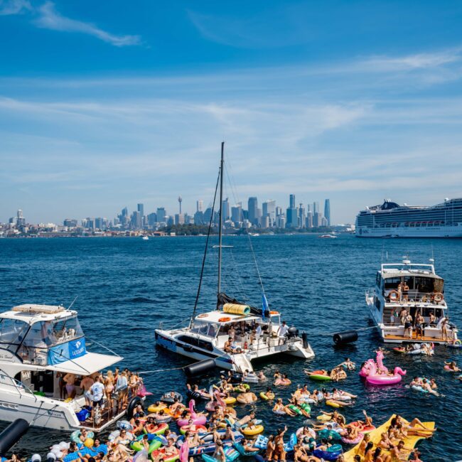 Crowd of people enjoying a sunny day on an urban waterfront, floating on colorful inflatables and surrounded by boats. City skyline is visible in the background.
