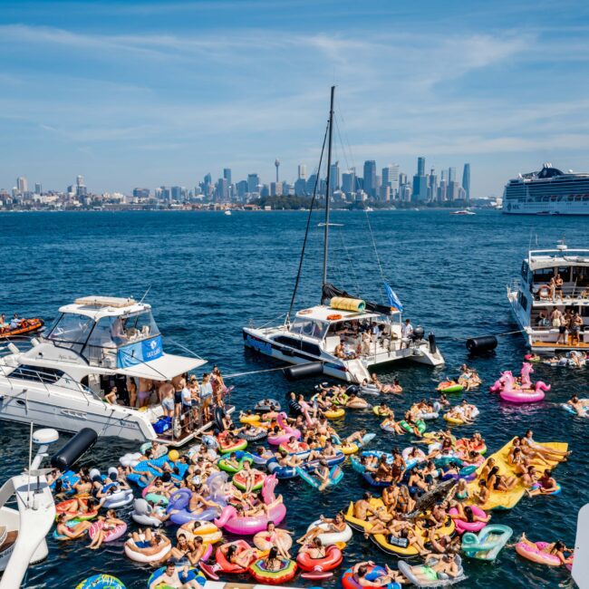 A lively scene of people on colorful inflatables gathered around boats on a sunny day. The city skyline and a large cruise ship are visible in the background, under a clear blue sky.