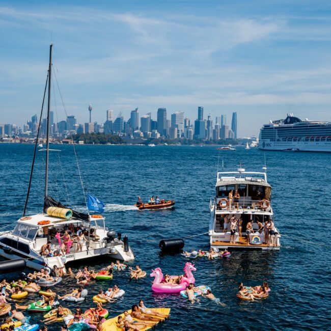 A lively scene on the water with people on inflatables and yachts, enjoying a sunny day. The city skyline, with tall buildings, is visible in the background. A large cruise ship is on the right.
