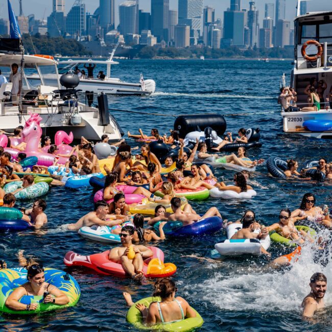 A lively gathering of people on colorful inflatable floats in a harbor, surrounded by boats. The skyline of a city with tall buildings is visible in the background under a clear blue sky.