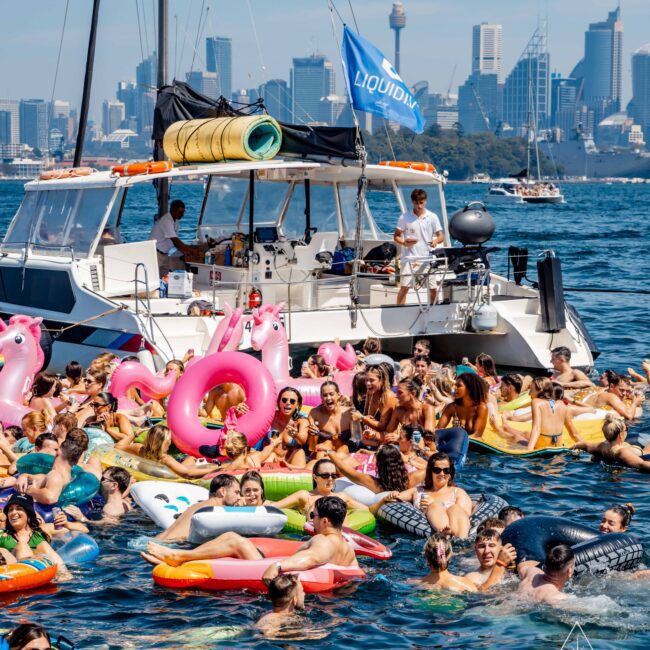 A large group of people enjoying a sunny day on colorful inflatable floaties near a boat in clear blue water. The city skyline is visible in the background, with tall buildings and a distinct tower. A blue flag on the boat reads "LIQUIDLY.