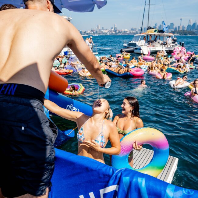 People enjoying a sunny day on the water. A person on a boat is pouring a drink for a laughing woman in a bikini holding an inflatable rainbow ring. Other people on floaters and boats are in the background.