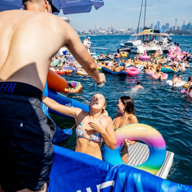 A person on a boat pours liquid into a smiling woman's mouth while she floats in the water holding a rainbow inflatable ring. Other people enjoy the water around them, with boats and a city skyline in the background.