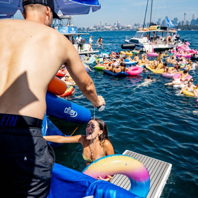 A man standing on a yacht sprays water on a woman in a pool, who is holding a colorful inflatable ring. Other people float on inflatables in the water nearby. Boats and a city skyline are visible in the background.
