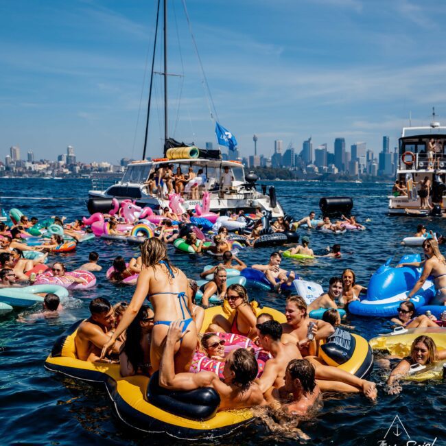 A crowded water party with people on inflatable rafts and pool floats in a harbor. Boats are moored nearby, and a city skyline is visible in the background. The scene is lively with colorful floats and festive atmosphere.