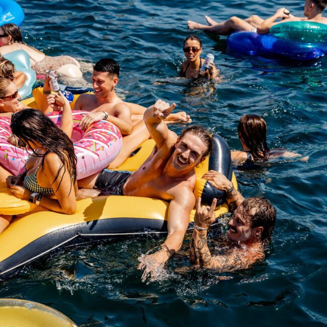 A group of people enjoying a sunny day in the water, surrounded by inflatable floats. Some are relaxing on the floats, while others swim nearby. Smiles and playful gestures are visible, creating a lively and fun atmosphere.