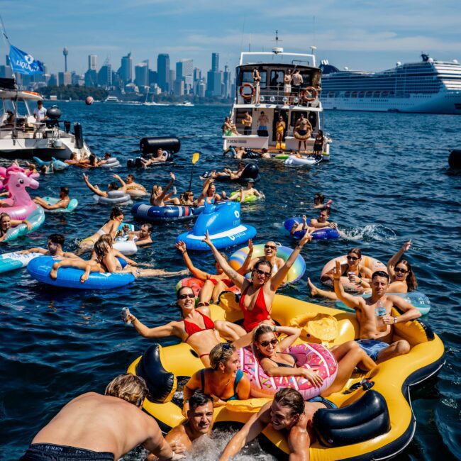 People are having a party on inflatable floats and swimming in a harbor with the city skyline in the background. A party boat is nearby, and the scene is lively and festive with bright sunshine and clear blue skies.
