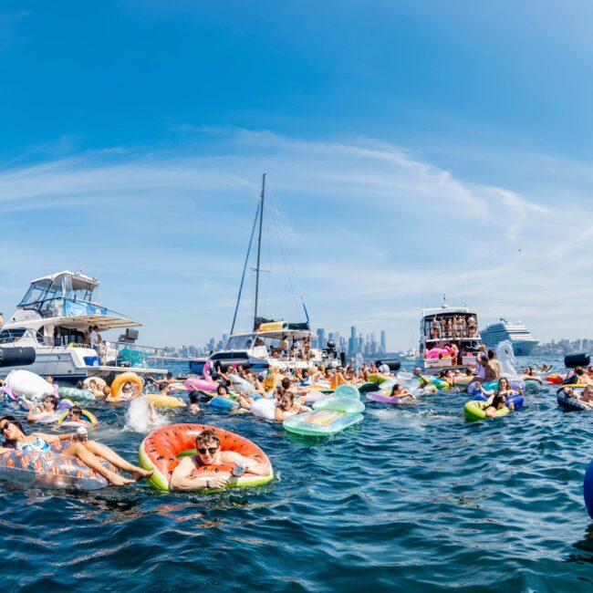 A group of people are on colorful inflatables in the water, enjoying a sunny day. Boats are nearby, and a cityscape is visible in the background under a blue sky with wispy clouds.
