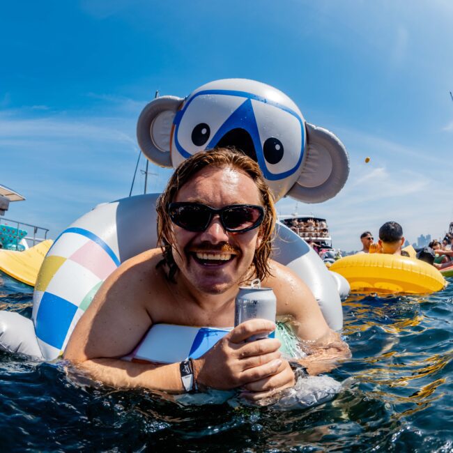 A person with sunglasses smiles while lounging on a koala-shaped float in a body of water, holding a can. Boats and other people on floats are visible in the background under a clear blue sky.