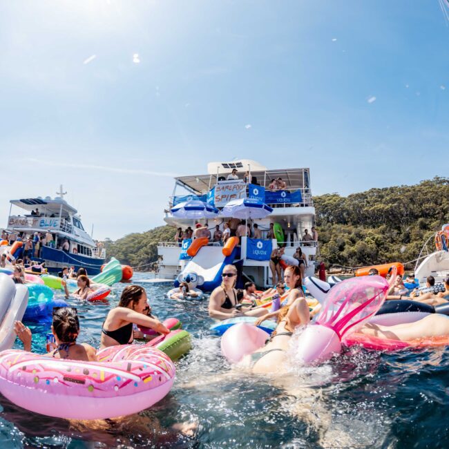 A lively scene of people enjoying a sunny day on the water, surrounded by inflatable floats in various shapes. Two large boats are anchored nearby, with a backdrop of lush greenery under a clear blue sky.