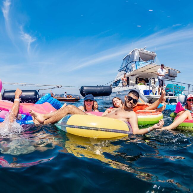 People relax on colorful inflatable rings in the water near a yacht. They are laughing and enjoying a sunny day. The scene includes a pink flamingo float and nearby boats, with a clear blue sky overhead.
