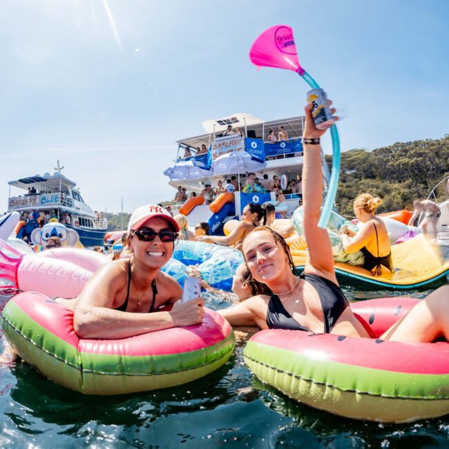 Two people in swimwear relaxing on colorful inflatable rafts in the water. One person is holding a drink and the other is holding up a drink pouch. Boats and other people on inflatables are in the background under a clear blue sky.