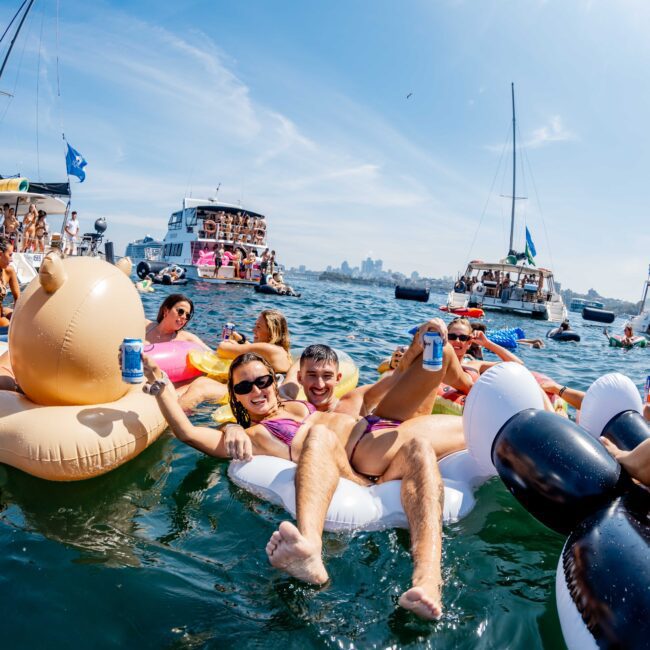 A group of people relax on colorful inflatable floats in a sunny bay, surrounded by yachts and boats. They are holding drinks and smiling. The water is calm, and the sky is clear, suggesting a festive and leisurely atmosphere.