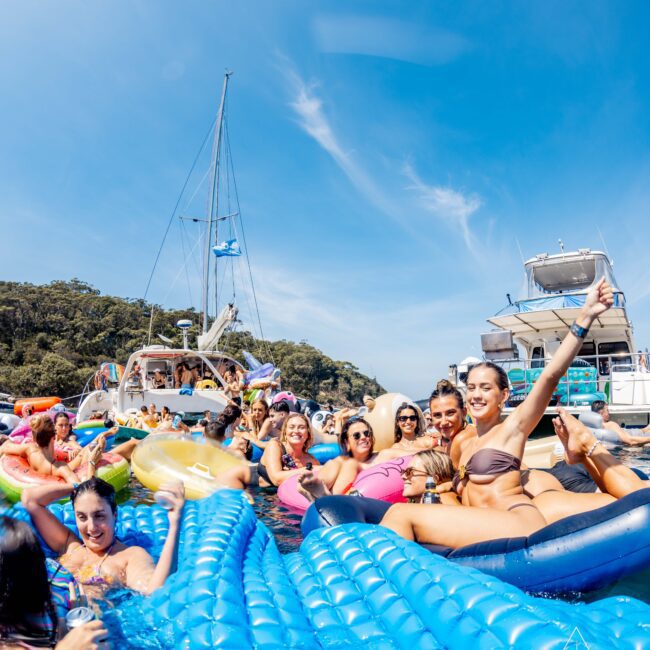 A group of people are lounging on inflatable rafts in the water near several boats under a clear blue sky. They appear to be enjoying a sunny day, with trees visible in the background. Some individuals are waving and smiling at the camera.