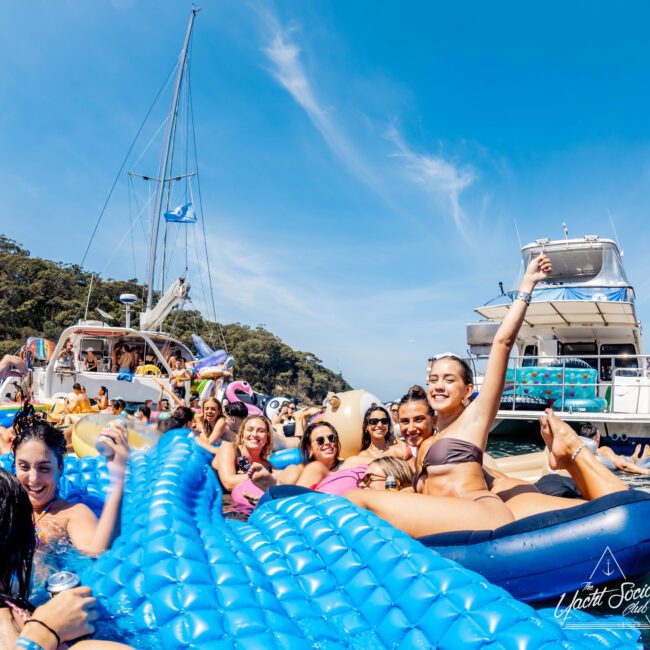 A group of people enjoying a sunny day on a yacht. They are lounging on blue inflatable floats in the water, smiling and posing for the camera. The water is clear, and other boats are visible in the background.