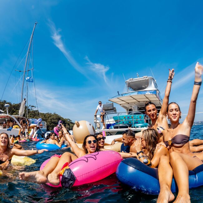 A group of people in swimwear are joyfully floating on inflatable rafts in the water next to a boat under a clear blue sky. They're raising their arms and smiling, surrounded by a lively and cheerful atmosphere.
