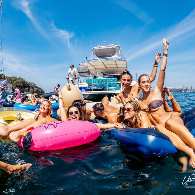 A group of people floating on colorful inflatables in the water, near a boat. They smile and pose for the camera under a sunny, clear sky. Other boats and a city skyline are visible in the background.