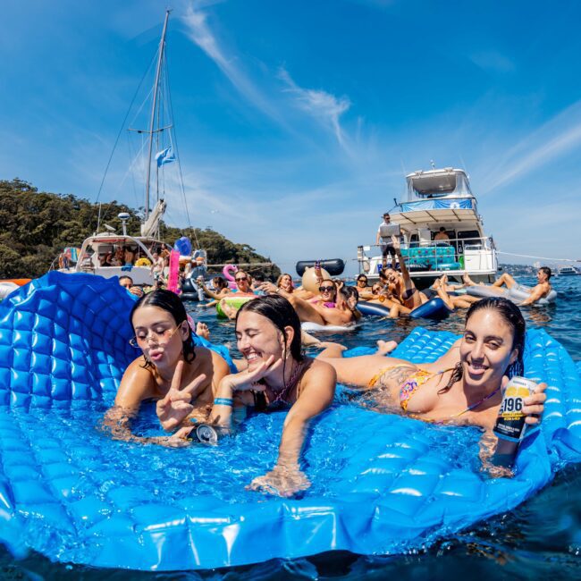 A group of people enjoying a sunny day on the water, lounging on a large blue inflatable raft. They are surrounded by boats, with others swimming and playing nearby, set against a backdrop of trees and a clear blue sky.