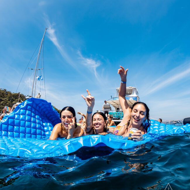 Three people enjoy a sunny day on a large blue float in the ocean, surrounded by boats. They are smiling, making hand gestures, and wearing swimwear. The sky is clear, and there are trees in the background.