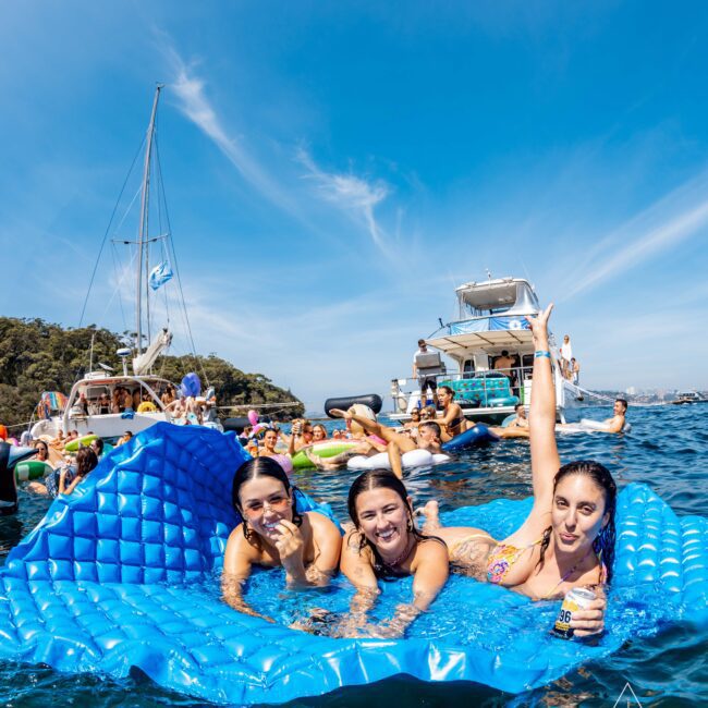 Three women lie on a blue inflatable mat in the water, smiling and posing. Behind them, a group of people relax on boats and inflatables under a clear blue sky, enjoying a day at sea.